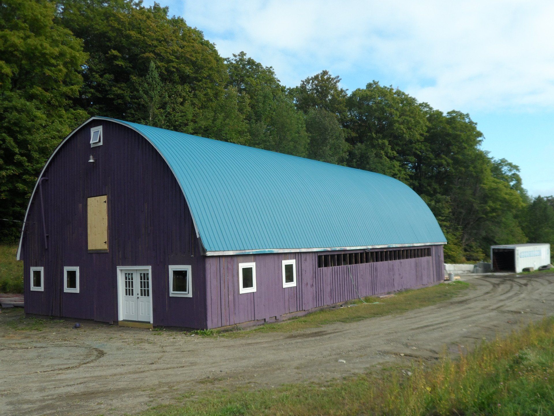 Purple barn with blue roof, white door and windows, dirt road, green trees in background.