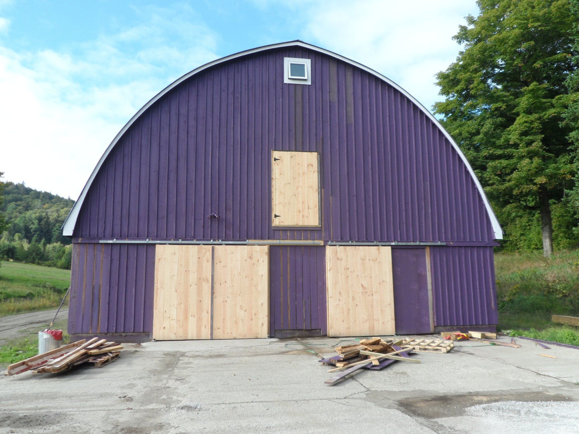 Purple Quonset barn with plywood doors and window on a concrete pad.  Wood piles rest on the ground.