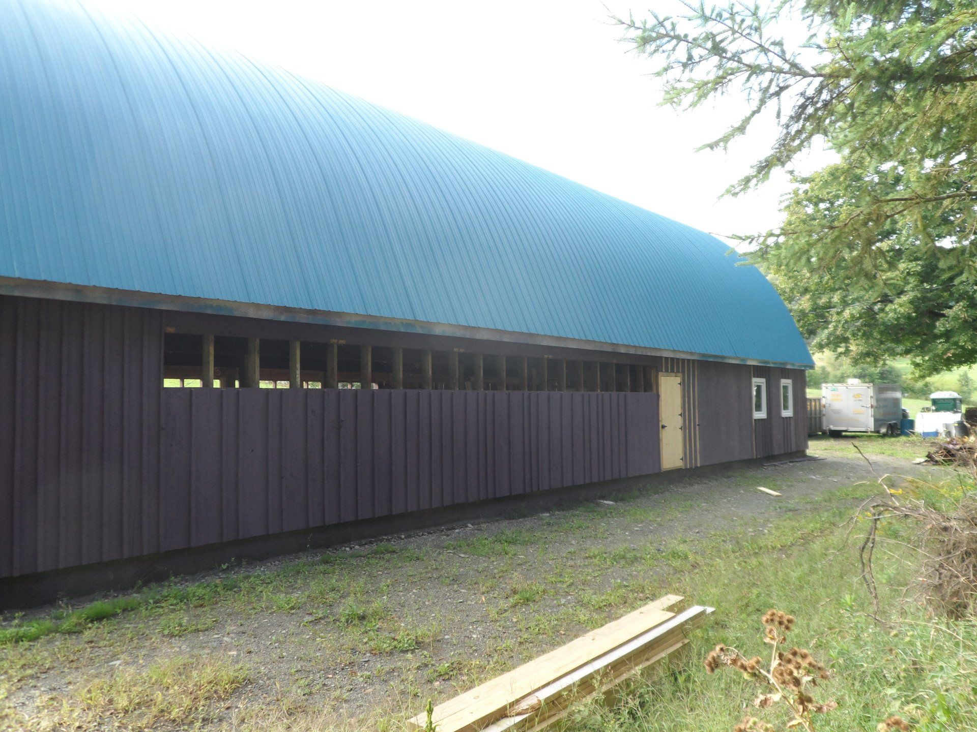 Dark brown barn with blue curved roof, set on gravel, trees and sky background.