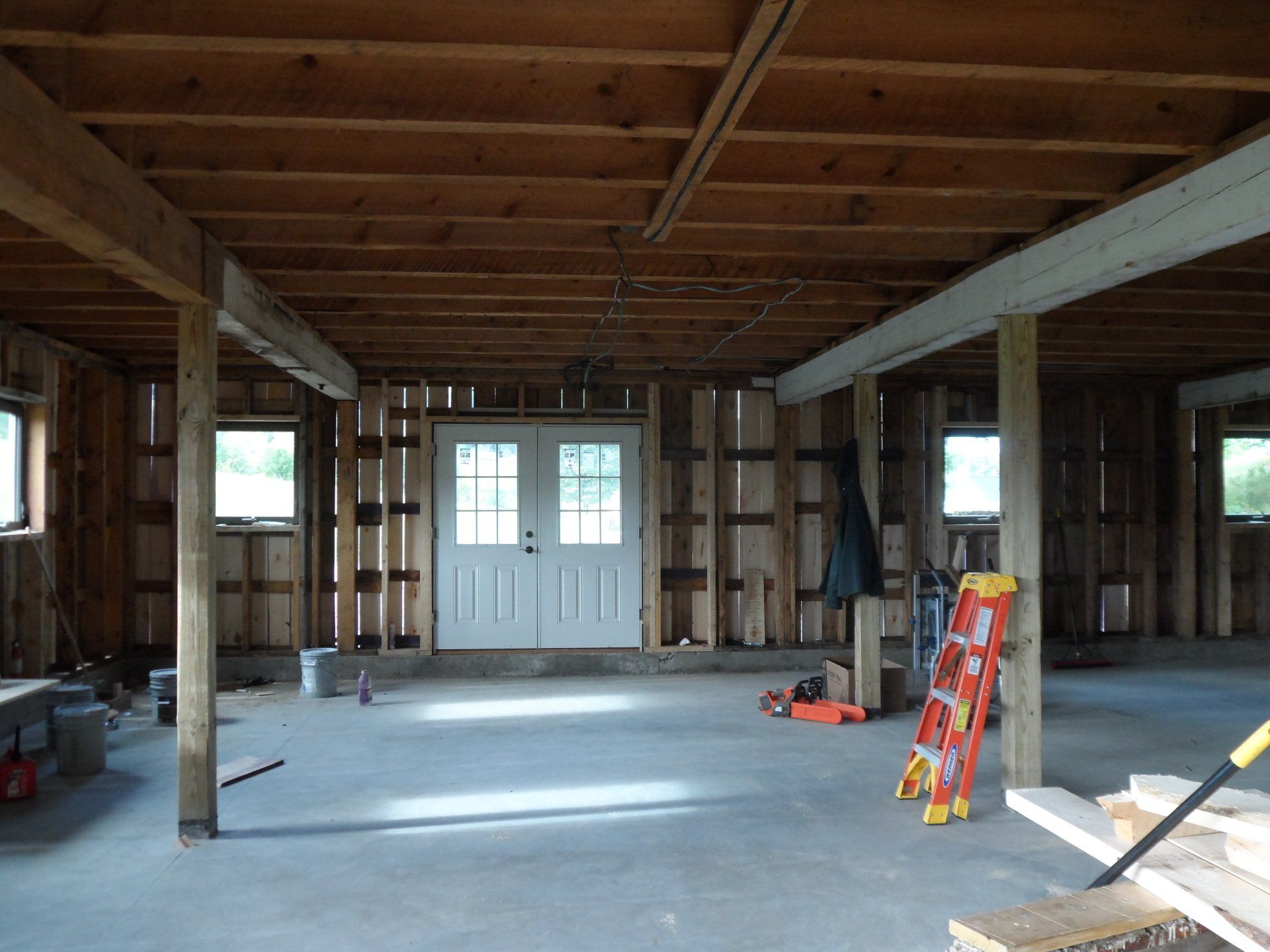 Interior of a building under construction, showing wooden beams, door, windows, concrete floor, and tools.