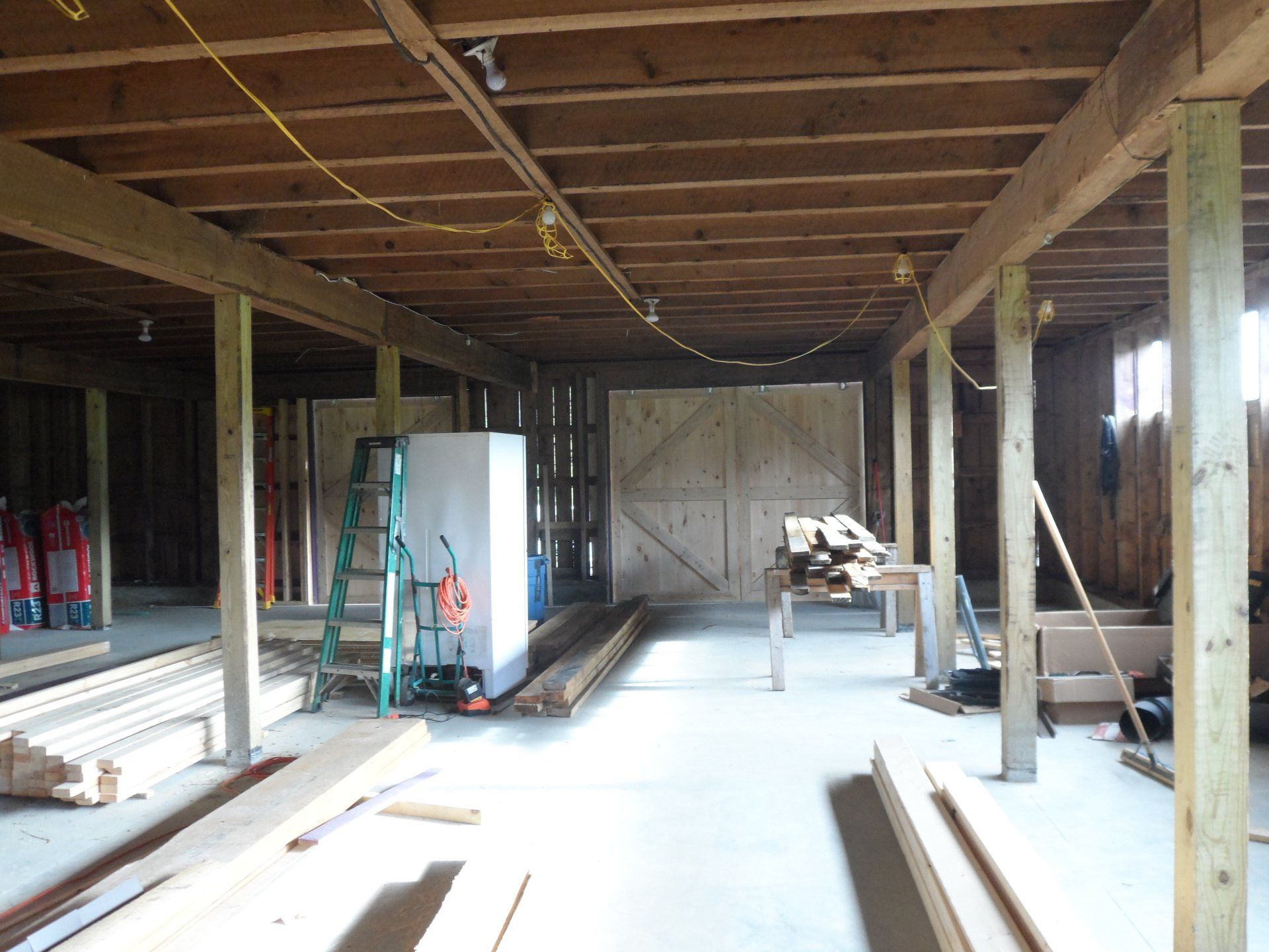 Interior of a barn under construction, with exposed wooden beams, lumber, and a closed wooden door.
