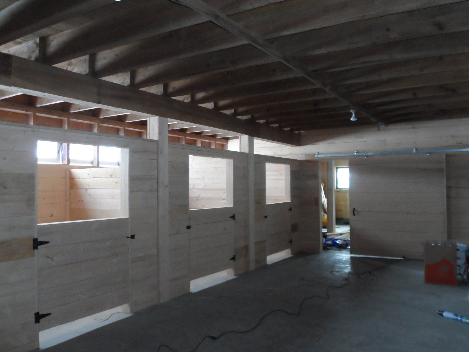 Interior view of a barn under construction with wooden stalls, open doorways, and a concrete floor.