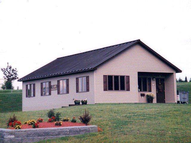 Single-story beige house with dark roof, front porch, and flower bed on a grassy lawn