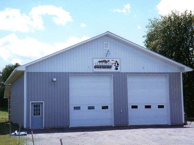 Gray metal garage with two white overhead doors and a small side door under a sign on the front.