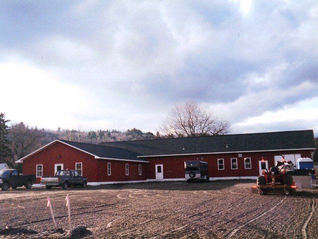 Red barn-style building on a gravel lot under a cloudy sky, with parked vehicles and equipment.