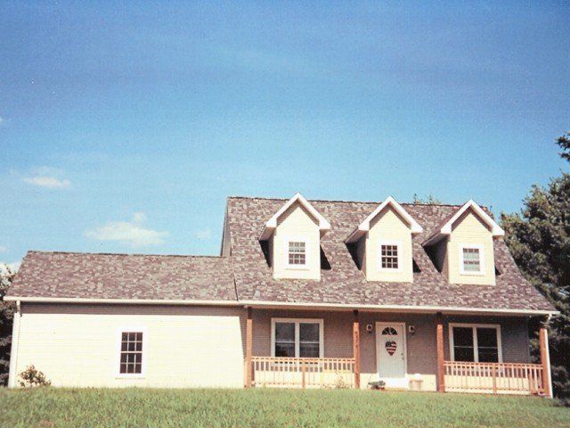 A one-story beige house with a brown shingled roof, three dormer windows, and a front porch, set under a clear blue sky.