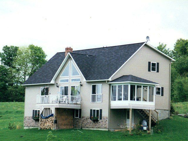 A two-story suburban house with a deck, a sunroom, and a wood-paneled basement area, surrounded by a grassy yard and trees.