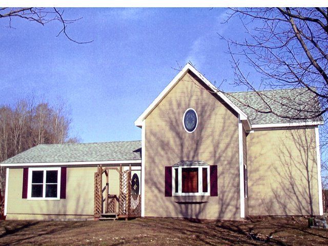 Small beige house with green roof, red shutters, and a front porch under bare trees.