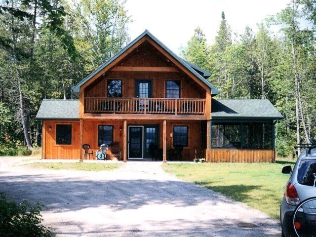 A two-story wood cabin with a balcony and screen porch, surrounded by trees with a gravel driveway in front.