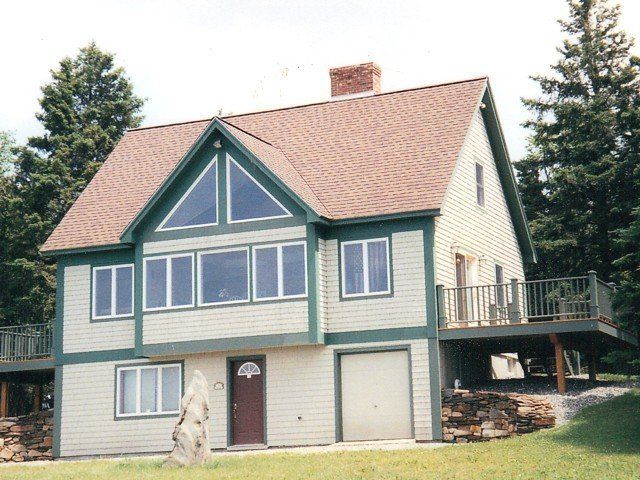 A beige two-story house with green trim, a small garage, a triangular front window, and a side deck nestled by pine trees.
