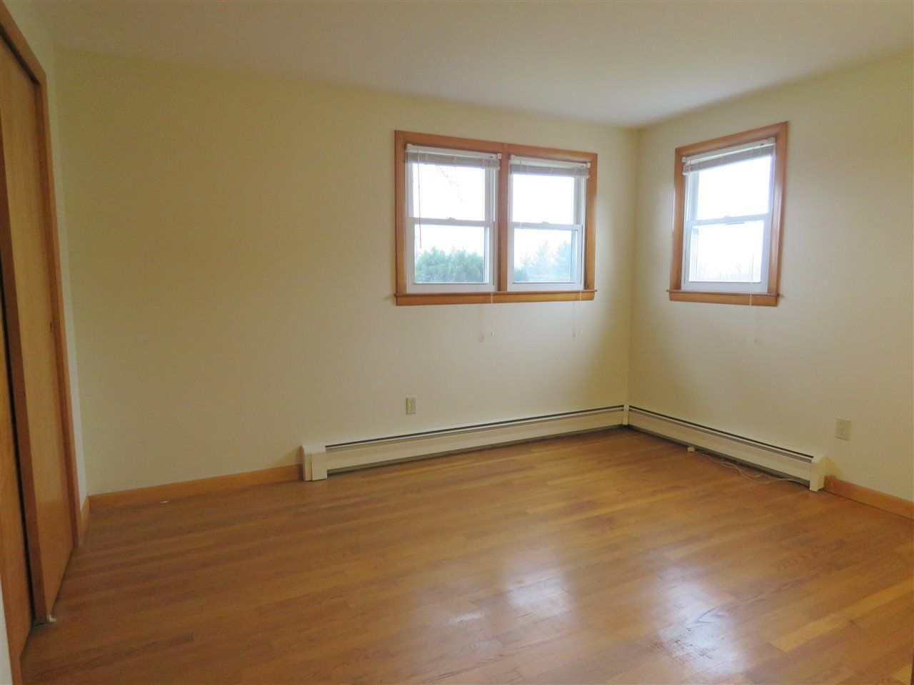 Empty room with hardwood floors, two windows, baseboard heating, and a closet.