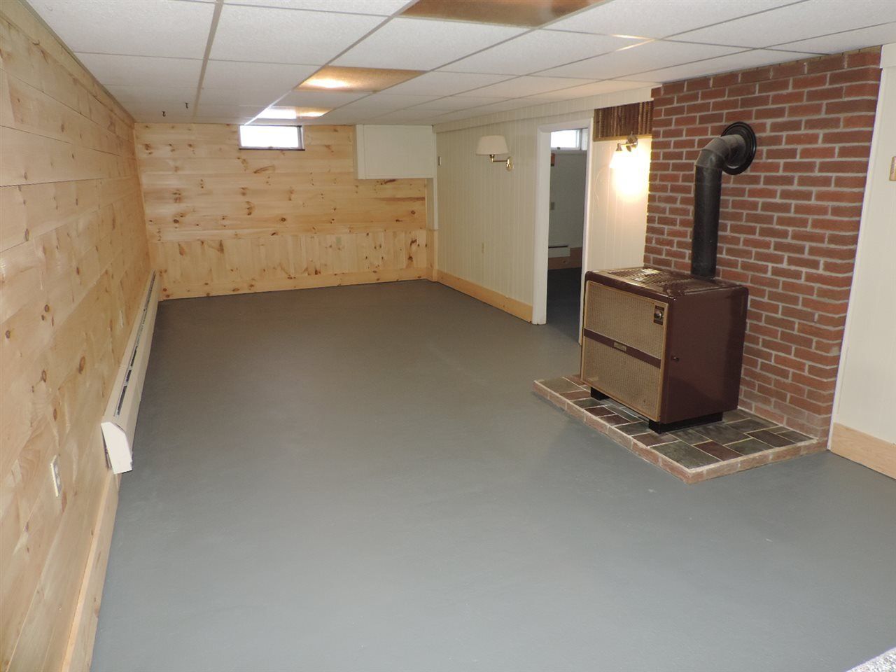 Basement with wood-paneled wall, brick chimney, gray floor, and a stove. The ceiling has a drop panel.
