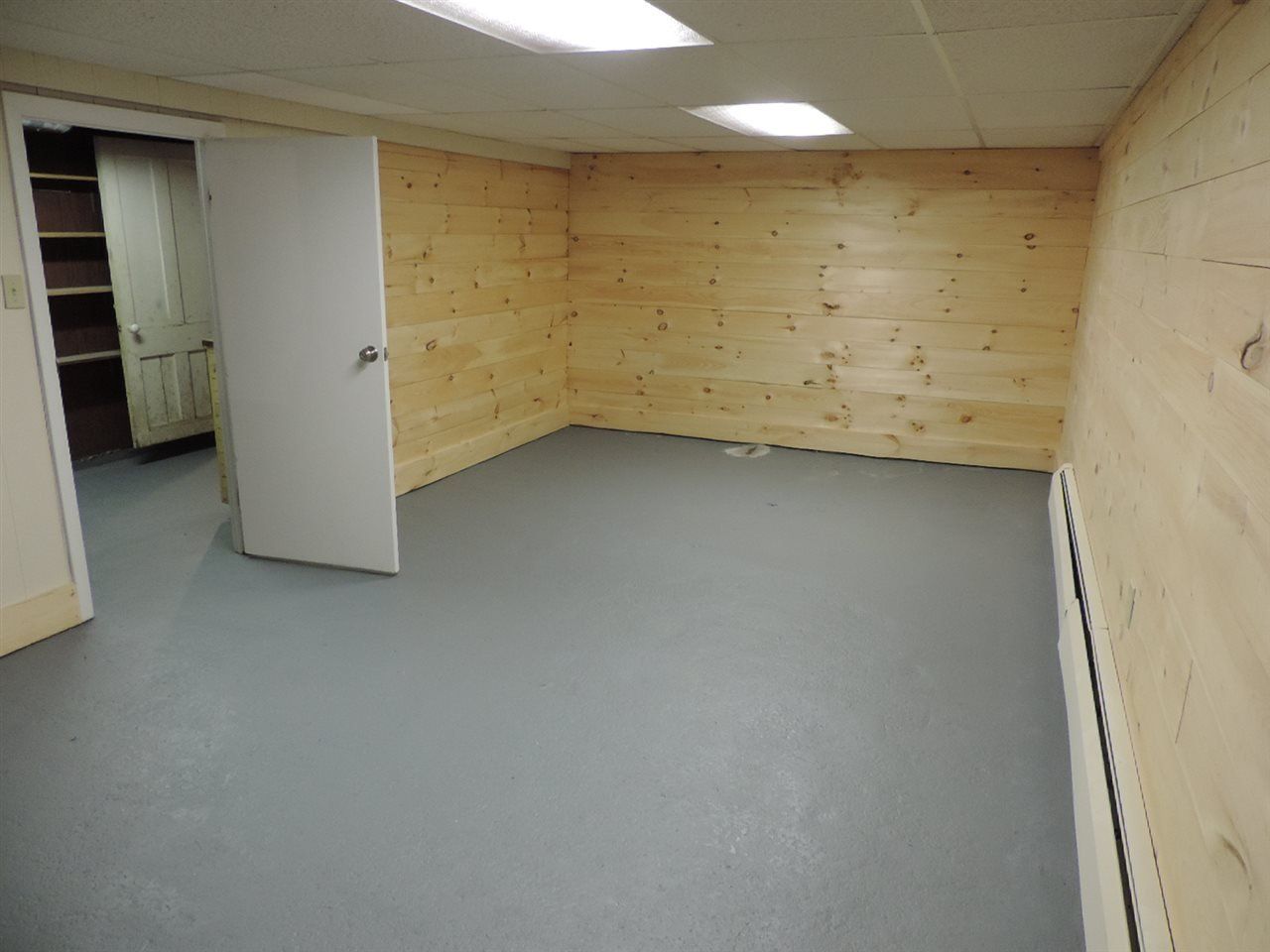 Empty room with gray floor and wood-paneled walls. White door and shelving visible.