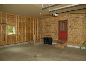 An unfinished garage interior with exposed wooden studs and walls, a red door, a window, and two black trash cans.