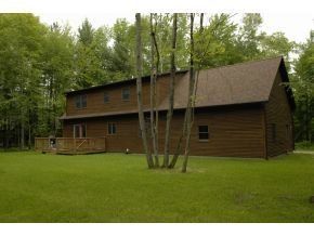 A two-story brown wood-sided cabin in a grassy, tree-filled clearing with a wooden deck extending from the side.