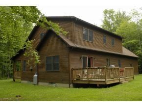 A brown, two-story wooden cabin sits in a grassy clearing surrounded by a dense forest, featuring a side wooden deck.