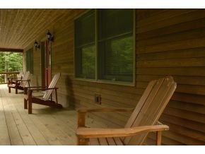 Wooden Adirondack chairs sit on a covered porch of a rustic log cabin, overlooking a wooded area.