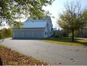 A white three-car garage and house with a long paved driveway, surrounded by trees on a sunny, autumn day.