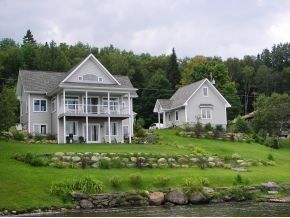 Two light-gray lakeside houses sit on a gentle, terraced grassy hill against a background of green trees.