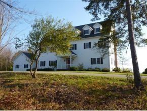 A large, white, multi-story house with dormer windows and a front porch, nestled among trees on a grassy hill.