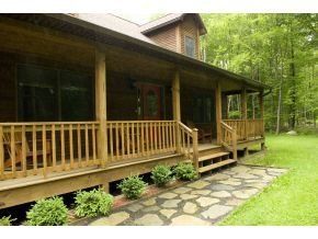 A wooden cabin with a wide front porch and railing, featuring a flagstone walkway surrounded by green bushes and trees.