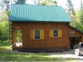 A wooden cabin with a green metal roof, shuttered windows, and an attached side garage, situated in a wooded area.