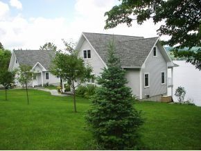Two light-gray houses with gabled roofs sit on a green lawn by a lake, with a spruce tree in the foreground.