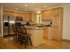 A kitchen featuring light wood cabinets, granite countertops, an island with three bar stools, and stainless steel fridge.