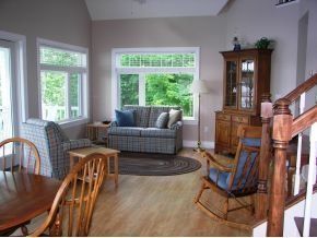 A sunlit living room with a plaid sofa, an armchair, a wooden rocking chair, a China cabinet, and a dining table.