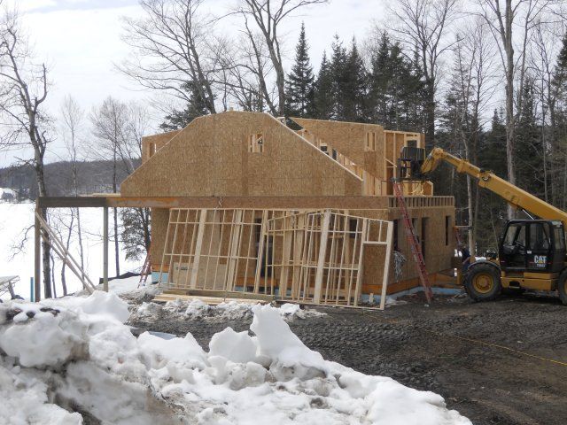 A construction site with a partially built wooden house frame and a yellow telehandler near snow-covered ground and trees.