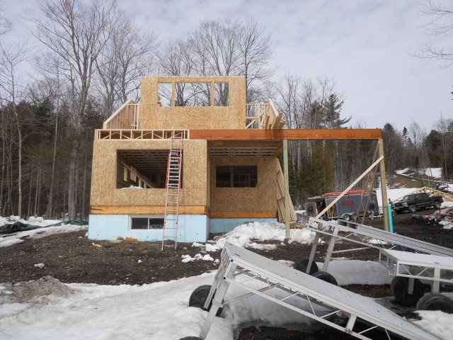A construction site showing a partially built wooden home frame with plywood walls over a light blue foundation in snow.