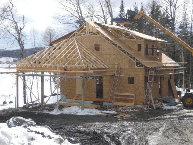 A construction site showing a wooden house frame being built in a snowy, rural landscape with a crane overhead.