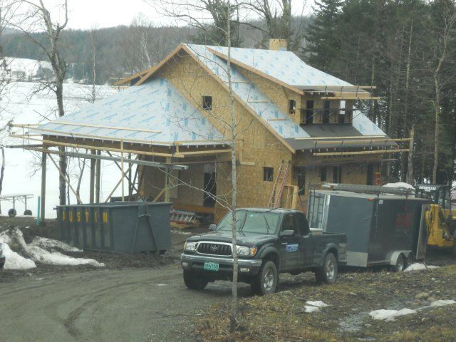 A house under construction with exposed wood framing, a partially covered roof, a dumpster, and a truck with a trailer.