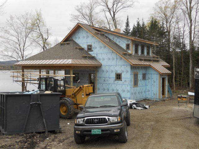 A house under construction with blue exterior insulation, a metal roof, a parked truck, and yellow heavy machinery nearby.