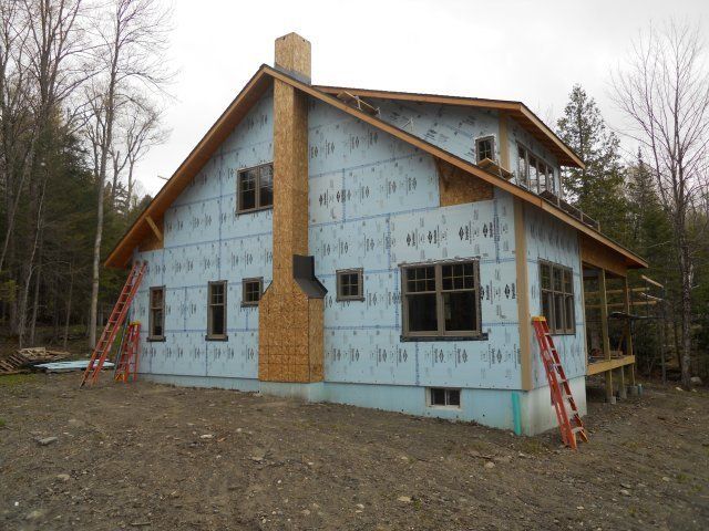 A house under construction with blue exterior foam insulation boards, a wooden chimney chase, and ladders on the sides.