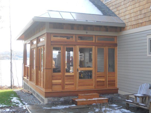 A modern wood-framed sunroom attached to a house with gray siding, overlooking a body of water with snow on the ground.