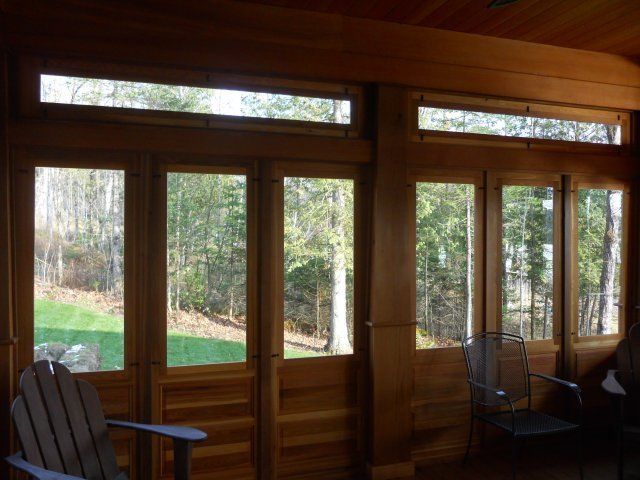 Wood-paneled sunroom with floor-to-ceiling windows and transom windows overlooking a wooded yard with two chairs.