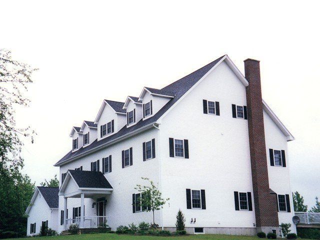 A large white, three-story house with a black roof, dark shutters, and a prominent brick chimney on the side.