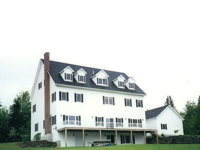 A three-story white house with a brick chimney, five dormers on the roof, and a large wooden deck in a grassy yard.
