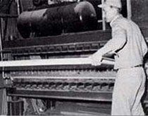 A person in uniform operates an industrial machine, feeding a flat board into a set of rollers.