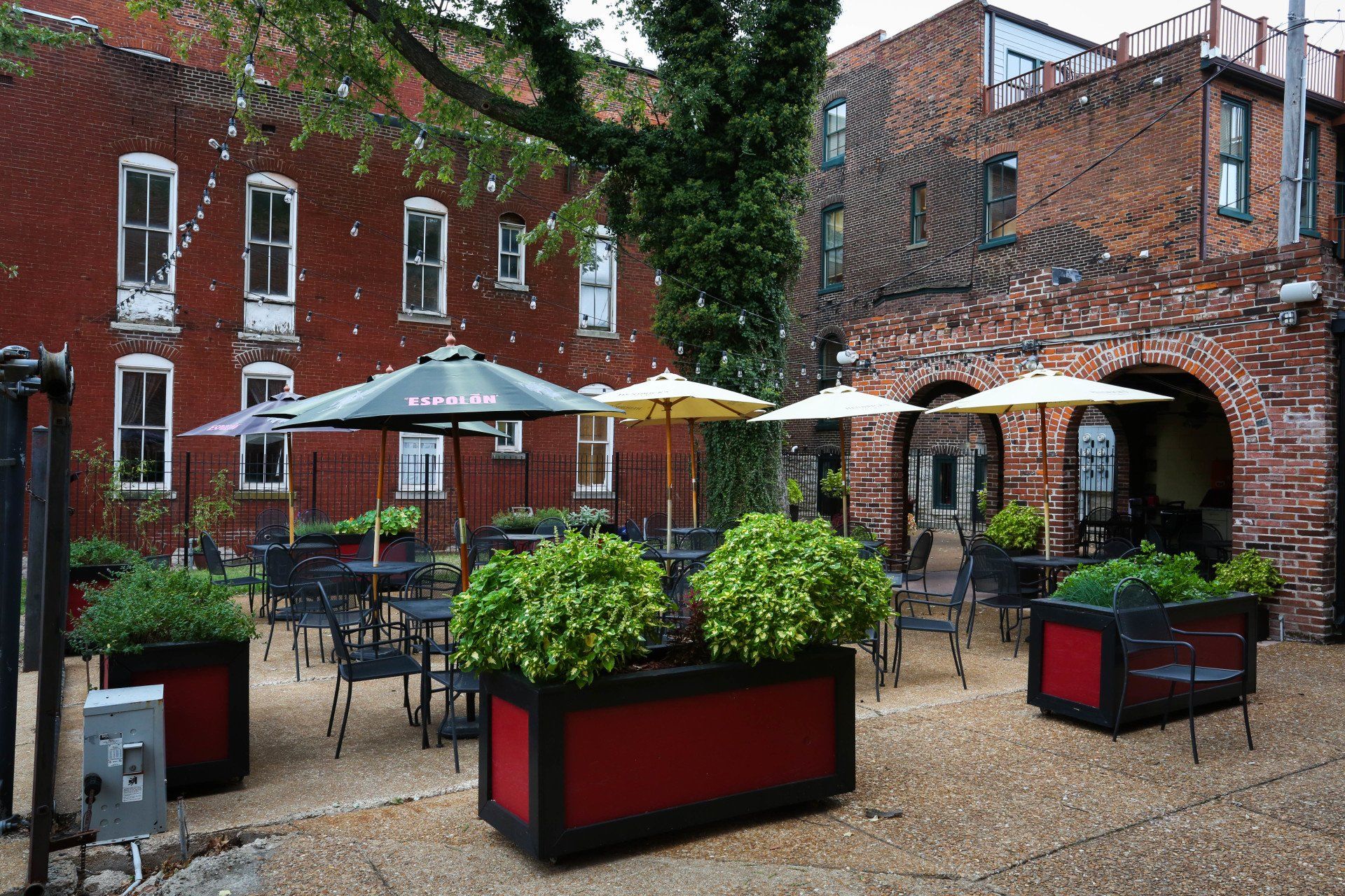 Outdoor brick courtyard with patio tables, umbrellas, and red planters under a tree