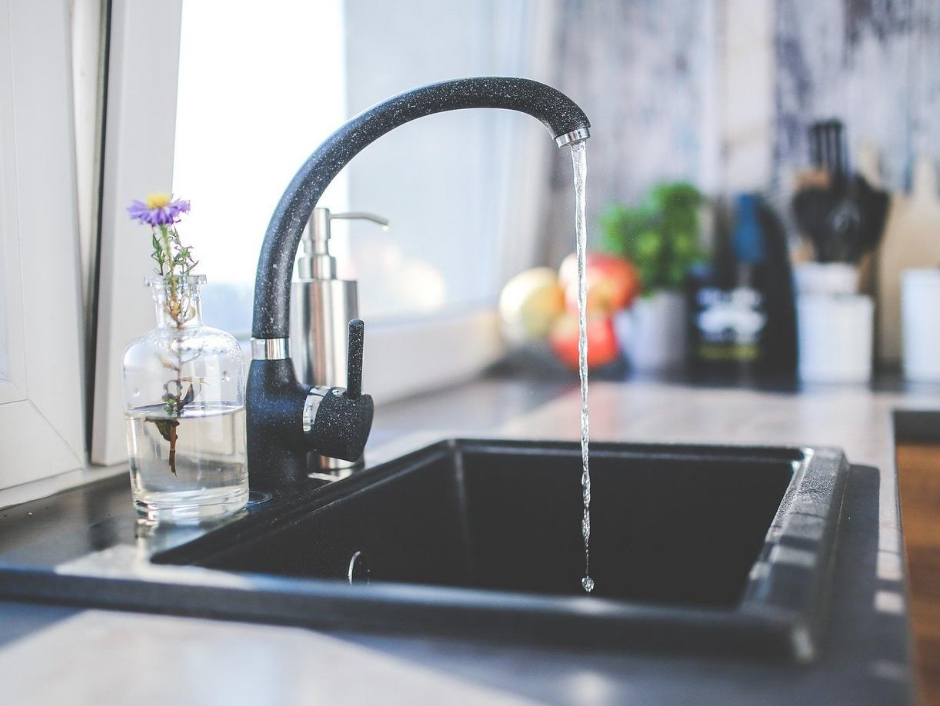 Black kitchen sink with running water, near a window with a vase of flowers.