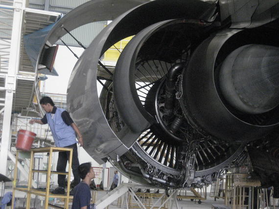 Workers inspecting a large jet engine in a hangar. One pours liquid from a bucket.