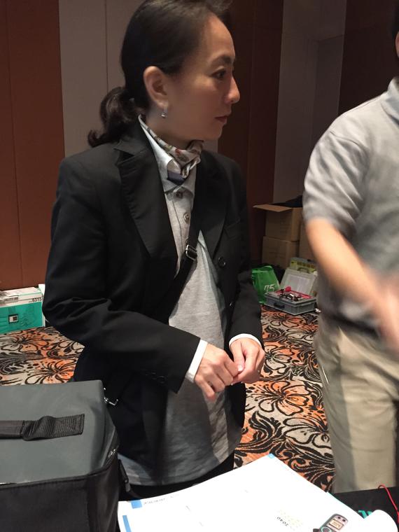 Woman in black blazer and gray shirt stands by a table; another person is blurred on the right.