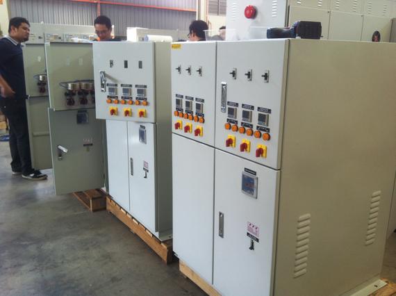 Workers inspecting electrical control panels in a warehouse; beige cabinets, yellow knobs, and digital displays.