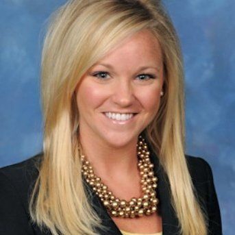 Headshot of a smiling blonde woman in a black blazer and gold necklace against a blue background