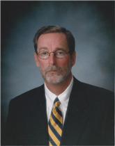 Studio portrait of a man in a dark suit, white shirt, and yellow striped tie, wearing glasses.