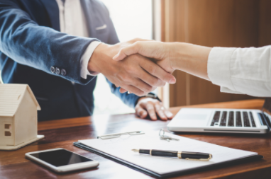 Two people shaking hands over a desk with a laptop, smartphone, house model, and paperwork