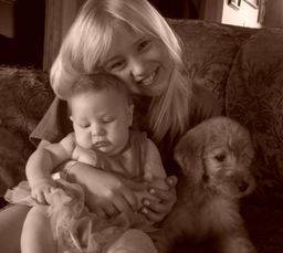 Young girl giving her baby sister a hug while snuggling with a goldendoodle puppy on the couch