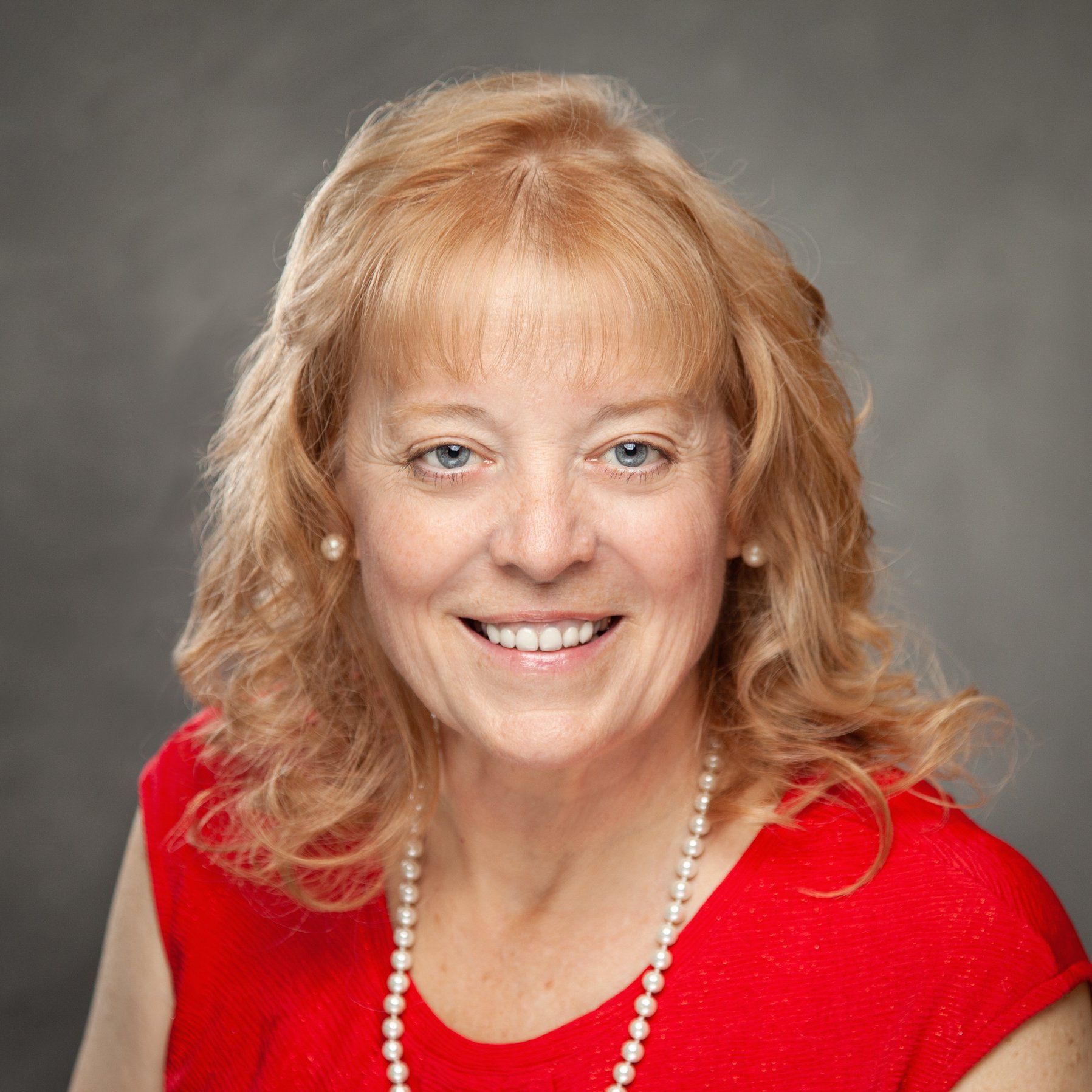 A woman wearing a red shirt and a pearl necklace is smiling for the camera.